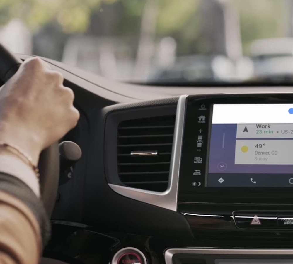 A woman hands on wheel driving across the city and the car front panel showing Hey Google turned on