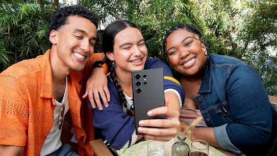 3 osoby uśmiechające się podczas robienia selfie. Osoba siedząca w środku robi zdjęcie telefonem z Androidem.