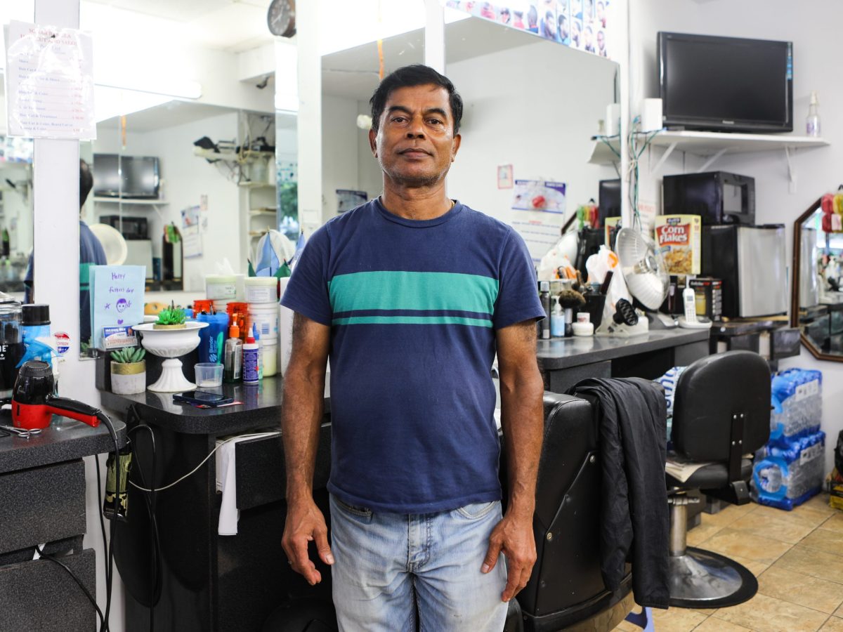Santi Shill stands in front of his barber stations with a large mirrors behind him. Shill is wearing a dark blue T-shirt with a turquoise stripe across the chest and blue jeans.