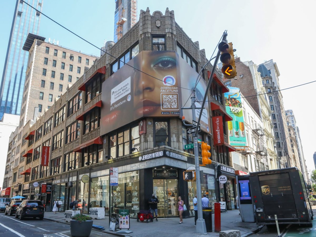 A corner building with a three sided billboard of a face up close. The eye of the poster faces out to the corner of 30th and 10th. In the background taller brick and glass buildings lead out of the frame.